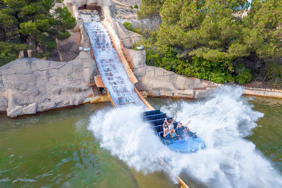 A water ride splashes as a blue boat with passengers descends from a slide into a pool, surrounded by trees and rocks.