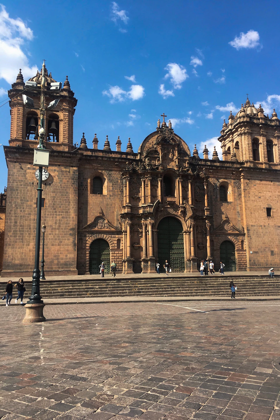 People walking in front of a historic church with twin bell towers under a blue sky with scattered clouds.