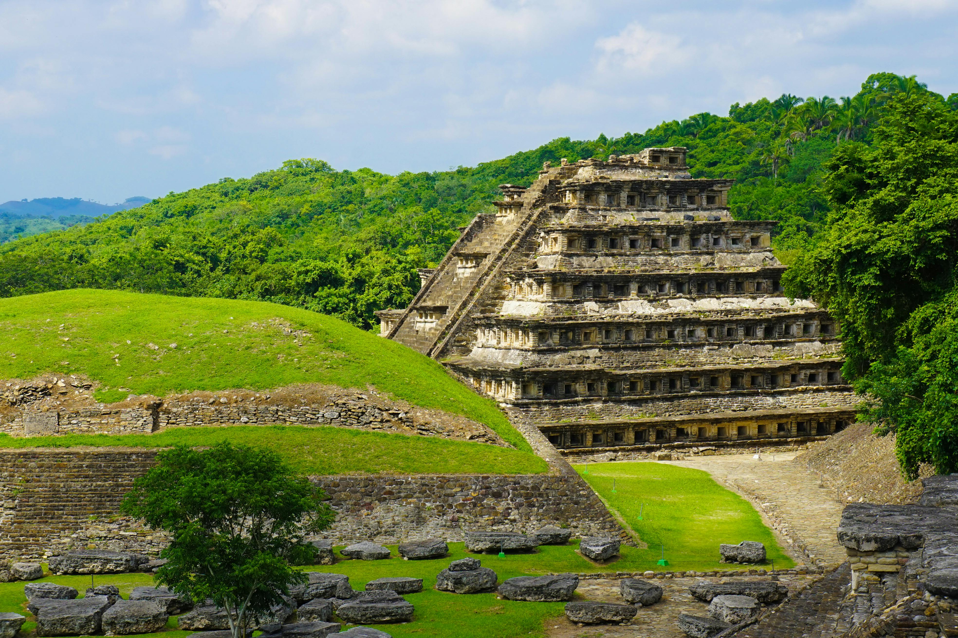 Eine mehrstöckige antike Pyramide steht in einer grünen Landschaft mit umliegenden Ruinen und dichtem Wald im Hintergrund.