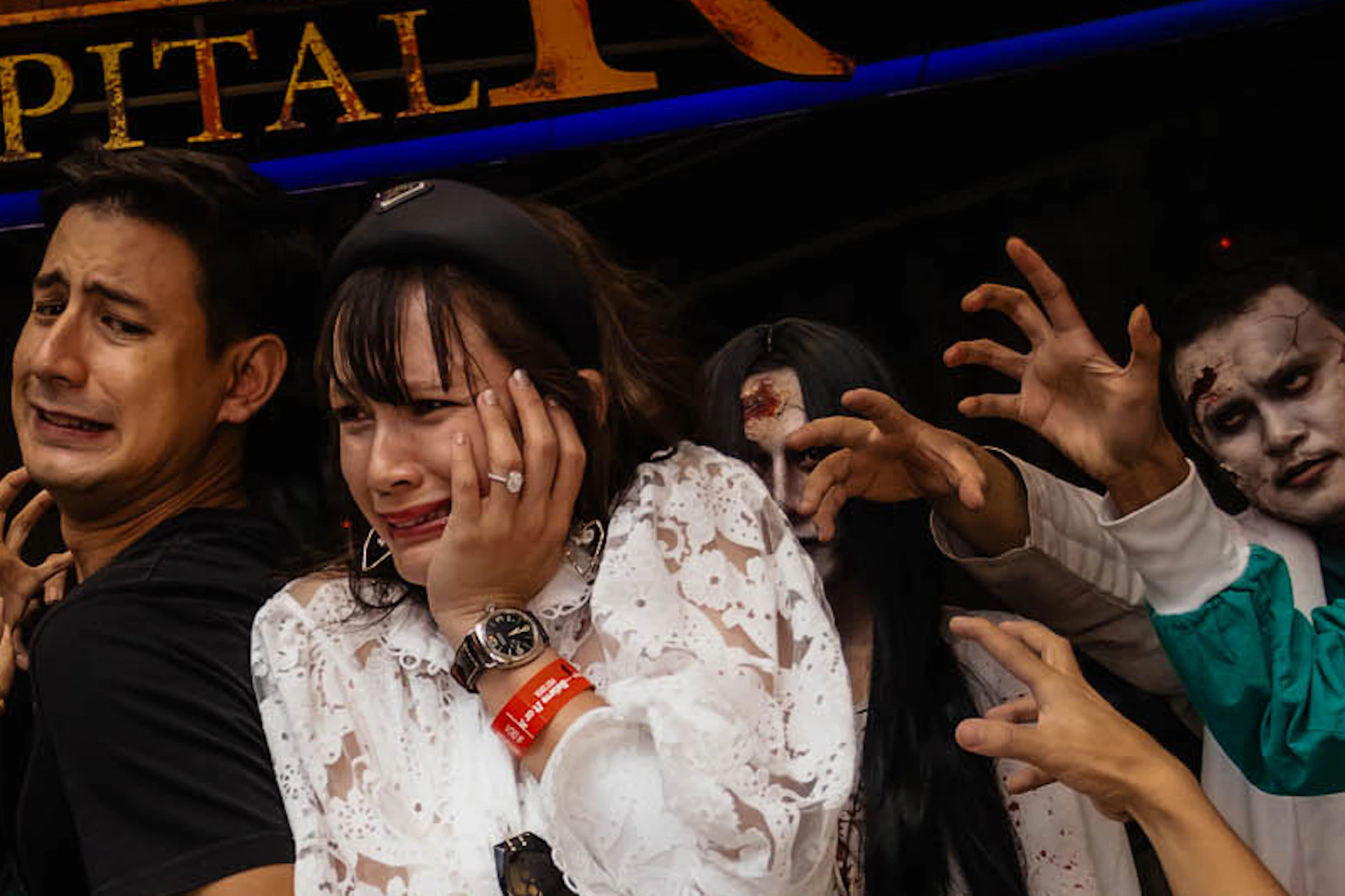 Two people appear frightened while surrounded by actors dressed as zombies in a haunted attraction labeled "Horror Hospital."