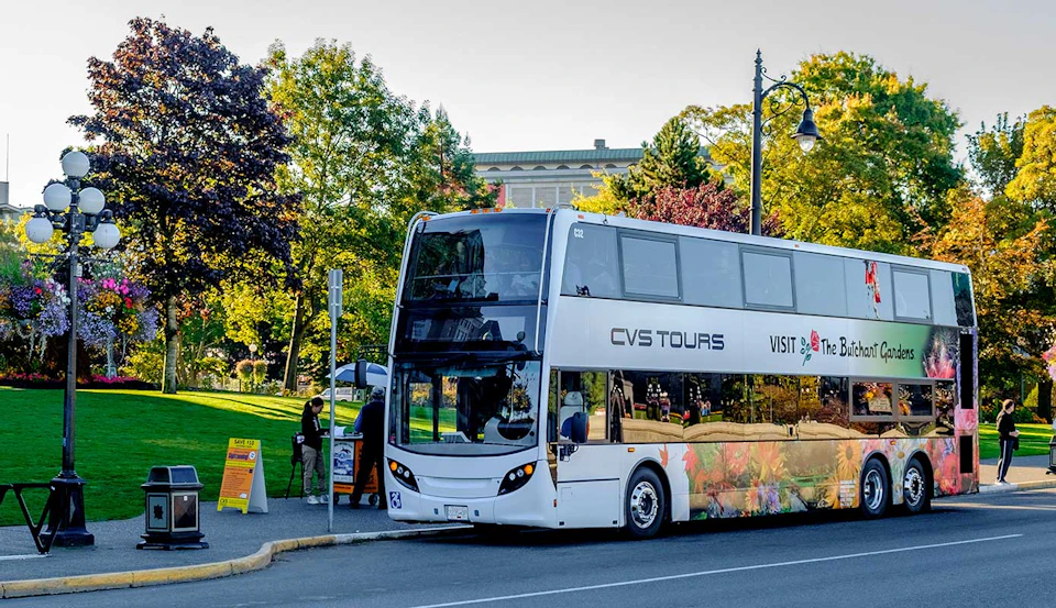 Double-decker tour bus parked near a park with lush trees and a building in the background. Several people are nearby.
