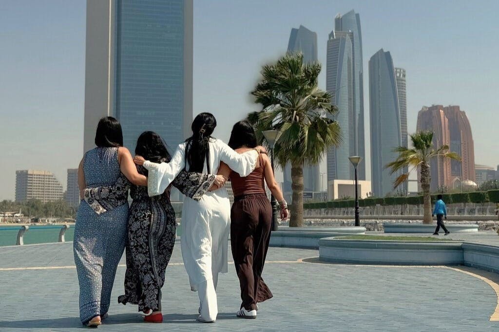 Four people with their arms around each other walk on a paved path, with modern skyscrapers and palm trees in the background.