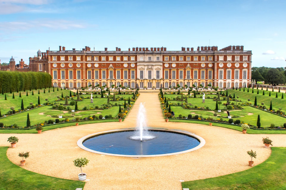 A grand red brick building with numerous windows overlooks a formal garden featuring a circular fountain and manicured lawns.