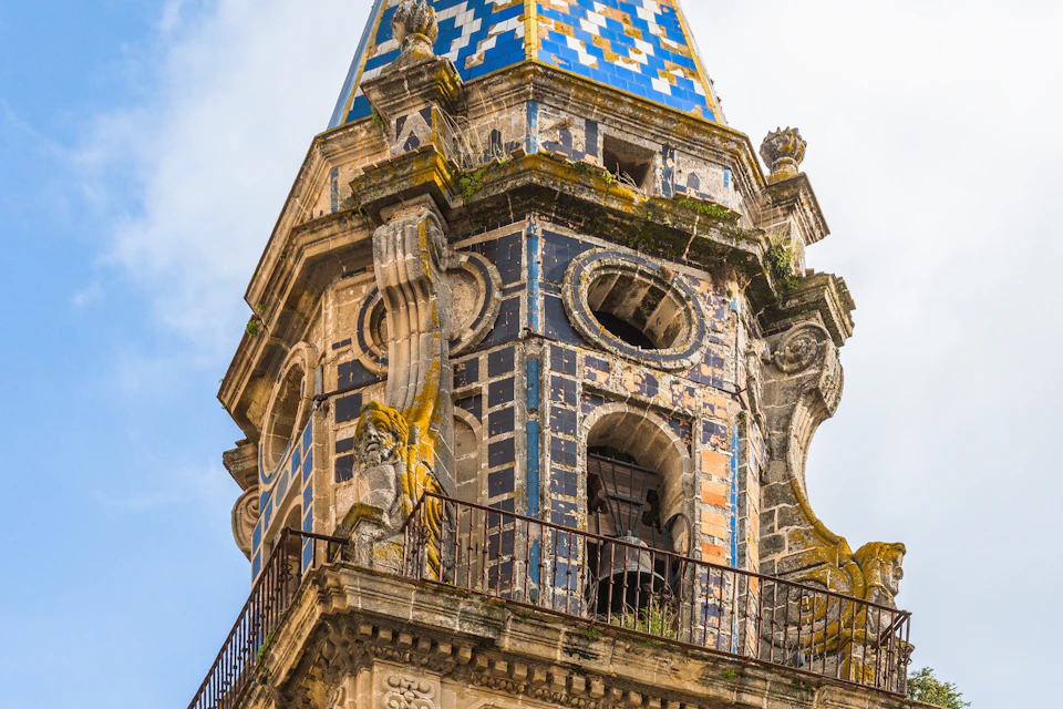 A close-up of a decorative tower with a blue and yellow tiled roof, intricate stonework, and moss-covered details.