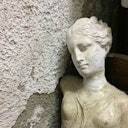 A weathered, classical-style bust leaning against a textured, peeling wall. The statue has a serene, downturned gaze.