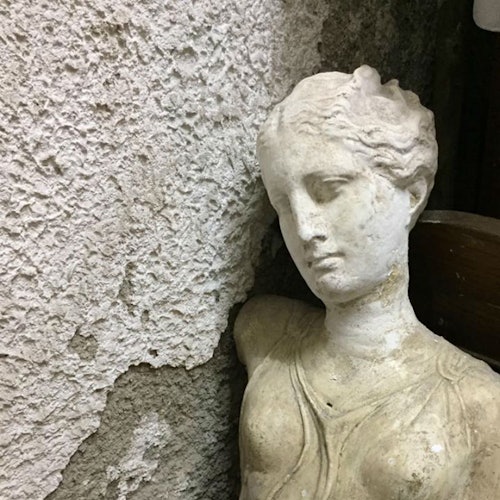 A weathered, classical-style bust leaning against a textured, peeling wall. The statue has a serene, downturned gaze.