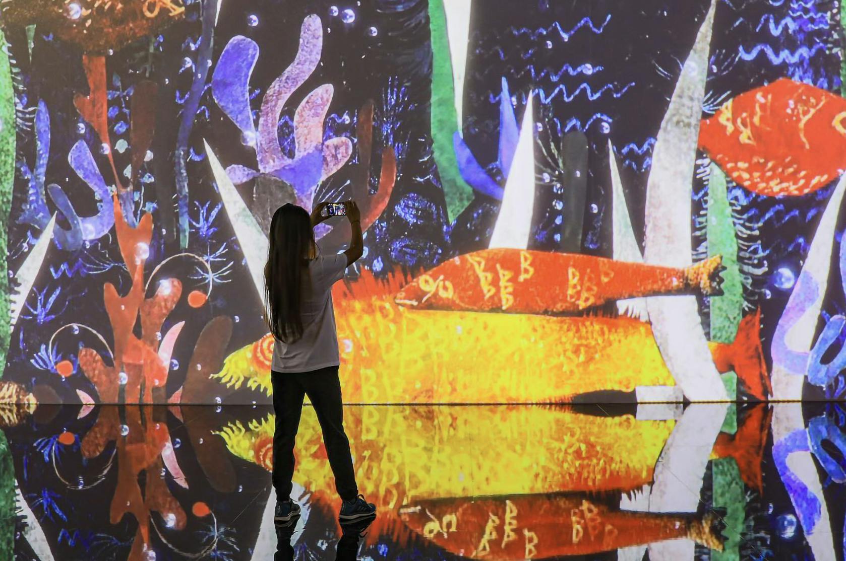Osoba stojí před barevnou digitální uměleckou projekcí a zachycuje ji pomocí smartphonu s odrazy na podlaze.