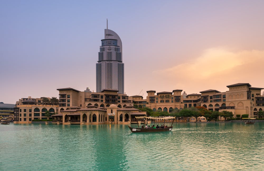 Um arranha-céus alto, com um topo curvo único, por detrás de edifícios de estilo tradicional à beira-mar; um barco com pessoas na água.
