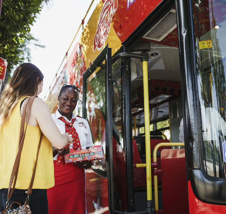 A woman in a yellow top boards a double-decker bus, greeted by a smiling conductor in a red and white uniform.