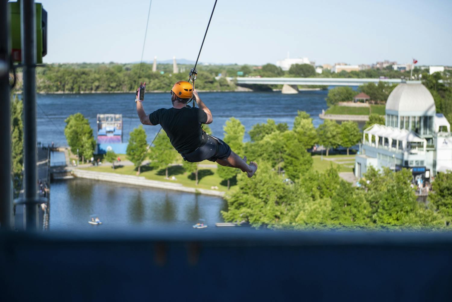 Persona con casco arancione che fa zip-lining su un fiume con alberi rigogliosi, ponti e strutture sullo sfondo.