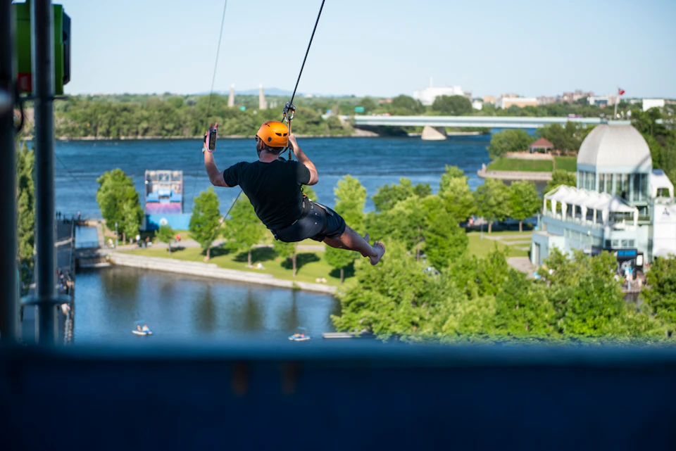 Person wearing an orange helmet zip-lining over a river with lush trees, bridges, and structures in the background.