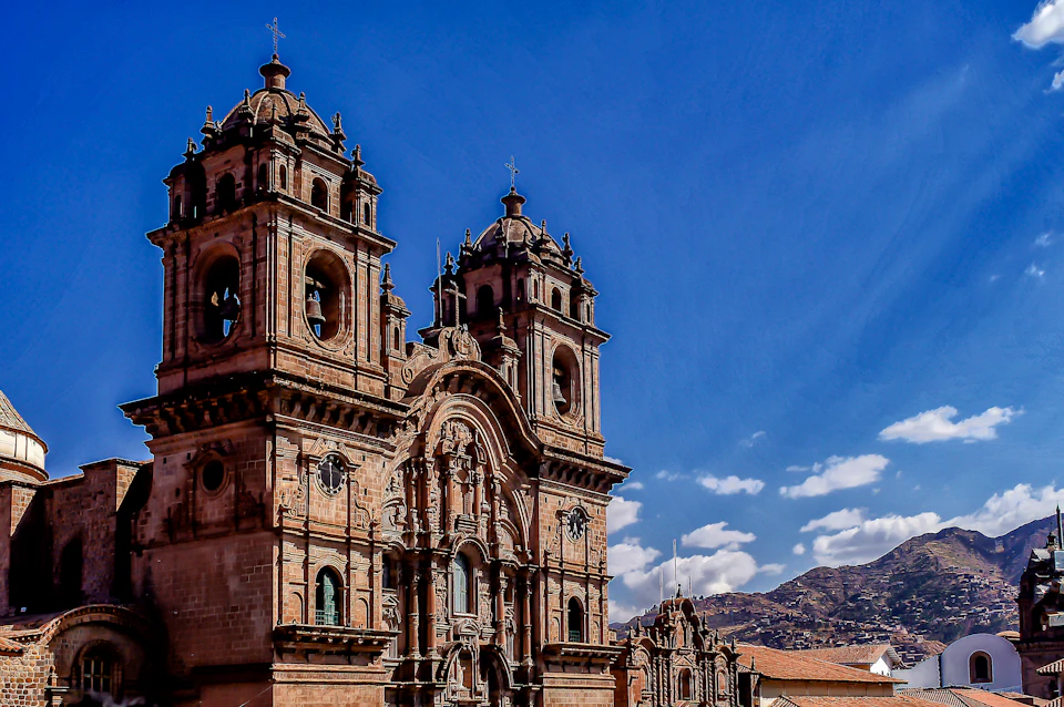 A historic stone church with two bell towers stands against a clear blue sky with mountains in the background.