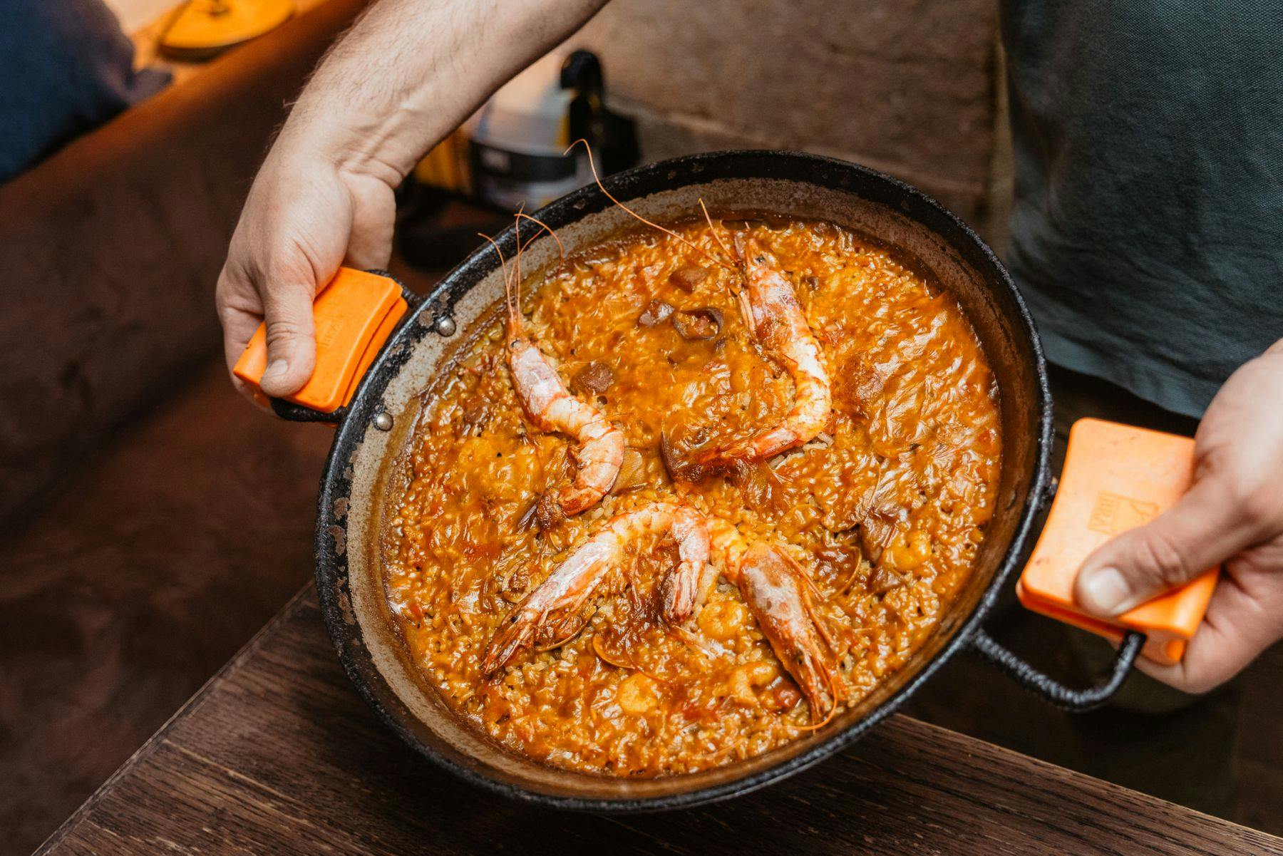 Uma pessoa segura uma panela de paella de marisco com camarão visível e arroz, apresentada numa superfície de madeira.