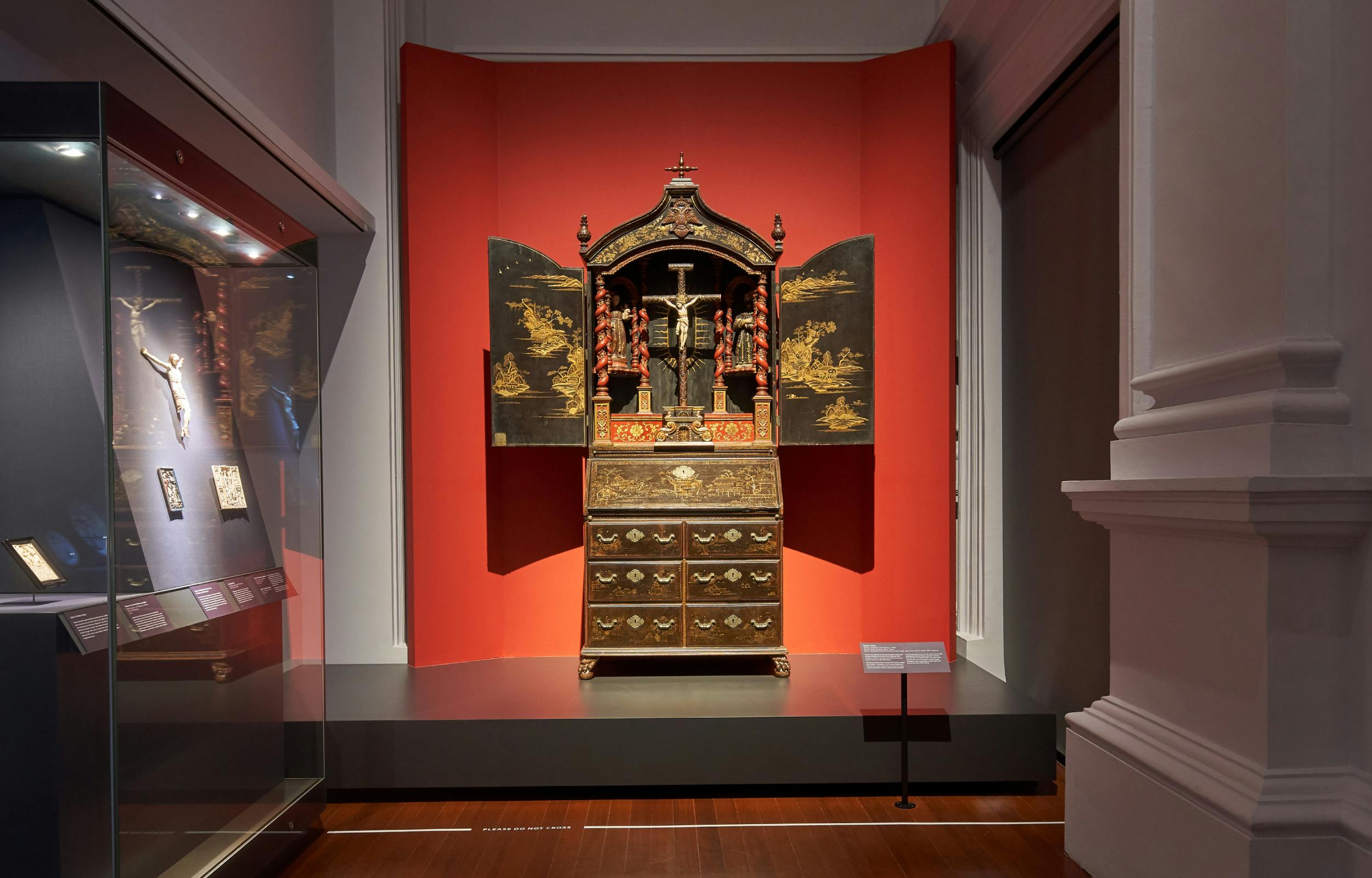 An ornate wooden cabinet with engraved doors open, revealing a crucifix and religious figures, displayed against a red background.