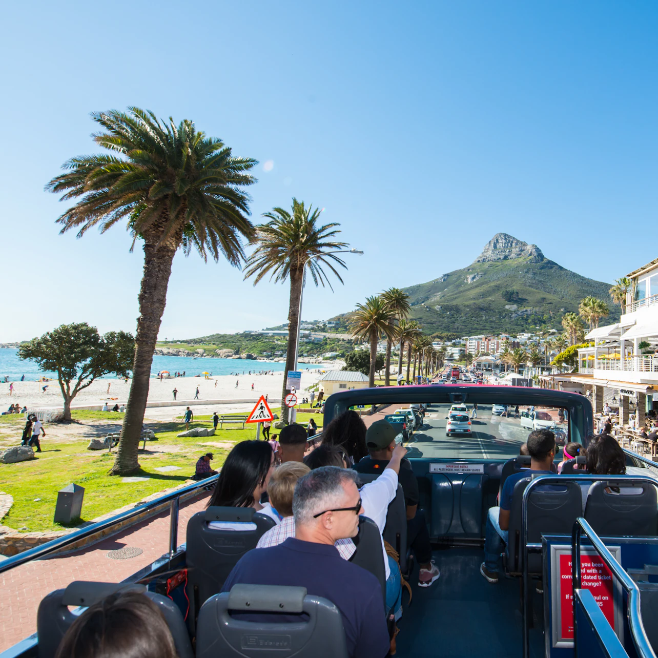 Tourists on an open-top bus touring a coastal town with palm trees, a beach, and a mountain in the background.