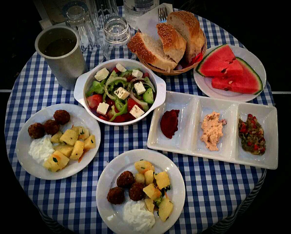 A table with a blue-checkered tablecloth holds plates of salad, bread, watermelon, small appetizer portions, and a drink.