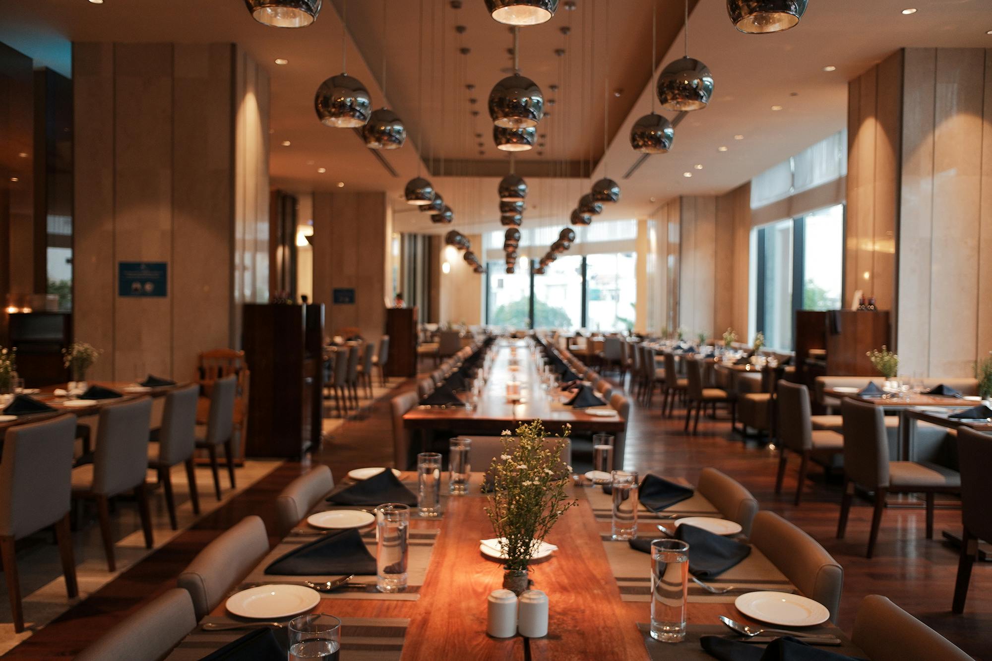 Przestronna, nowoczesna restauracja z długimi drewnianymi stolikami, zestawem do jadalni i dekoracyjnymi lampami wiszącymi.