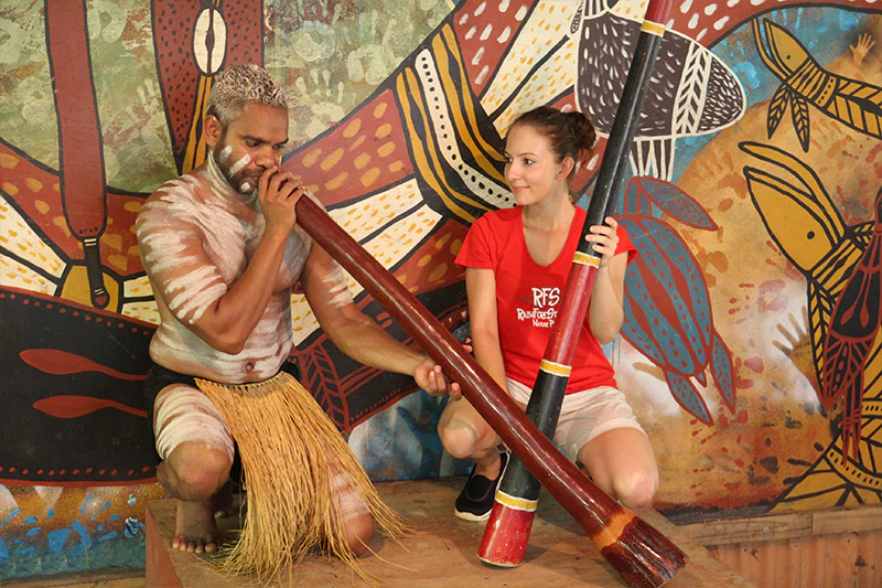 A person playing a didgeridoo while another person kneels beside them, holding a similar instrument, in front of an Indigenous art mural.