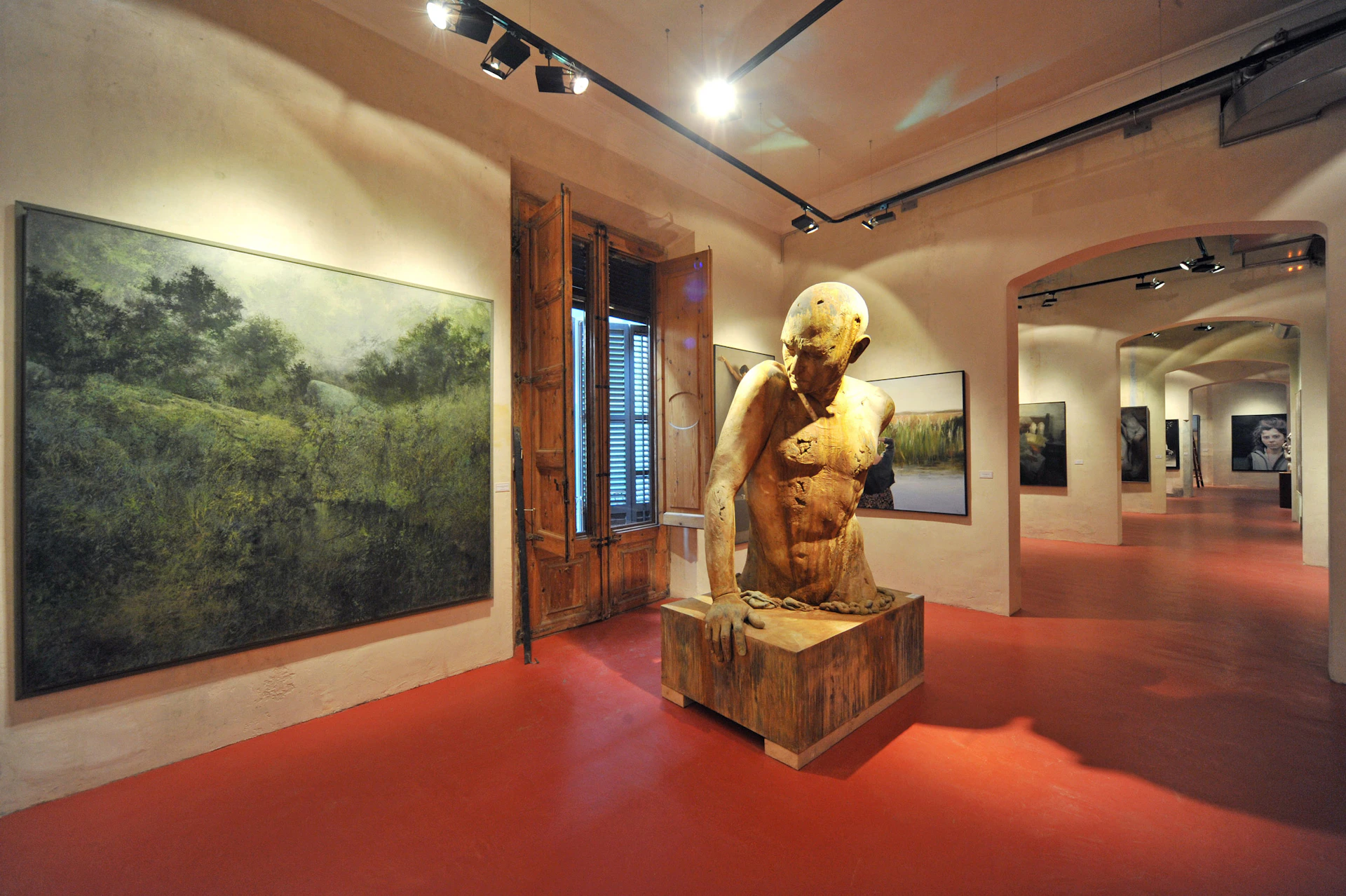 A gallery features a large wooden human sculpture, landscape paintings on the walls, and track lighting on a red floor.
