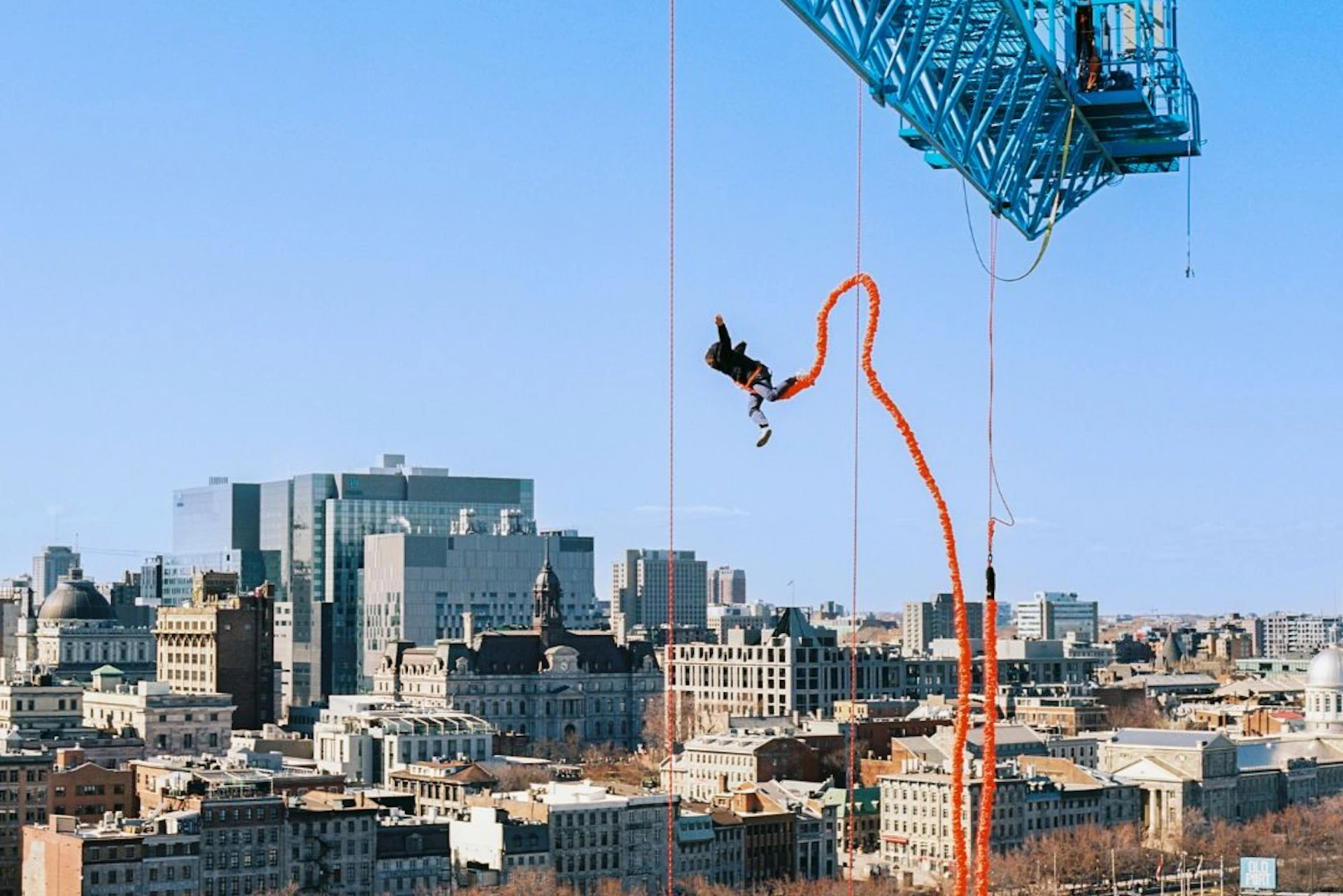 A person is bungee jumping from a large blue crane overlooking a cityscape with tall buildings on a clear day.