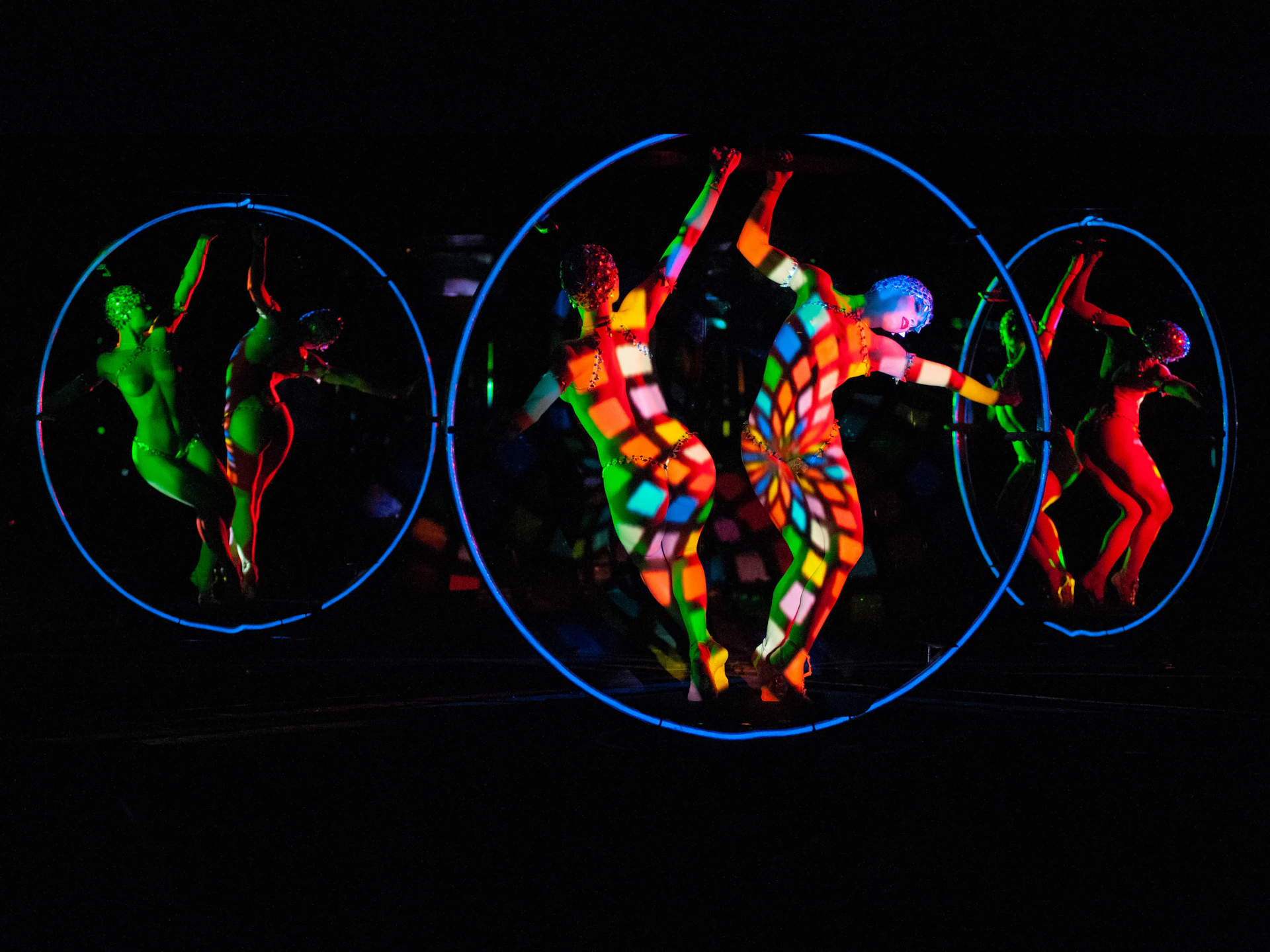 Performers in colorful bodysuits inside circular frames, with vibrant lights and patterns in a dark setting.