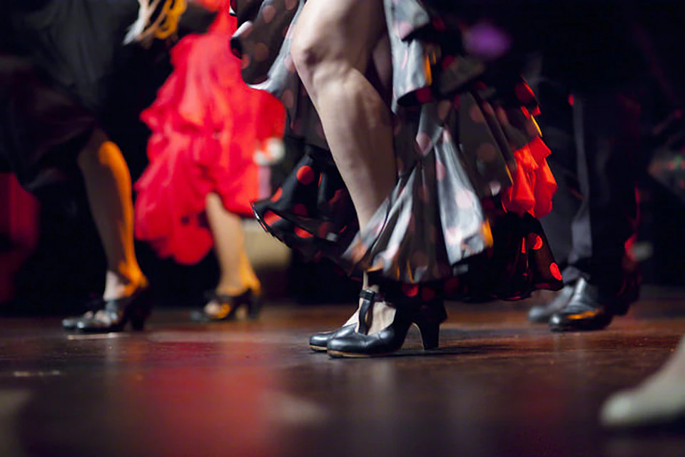 Dancers in colorful, ruffled dresses perform on stage, focusing on their legs and black dance shoes.