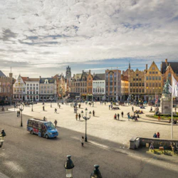 Ett stort offentligt torg med människor som går, färgglada historiska byggnader, ett monument och flaggor under en delvis molnig himmel.
