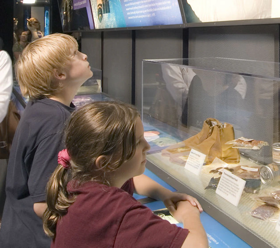 A woman and three children observe space exhibits and artifacts in a museum, with a satellite model displayed in the background.