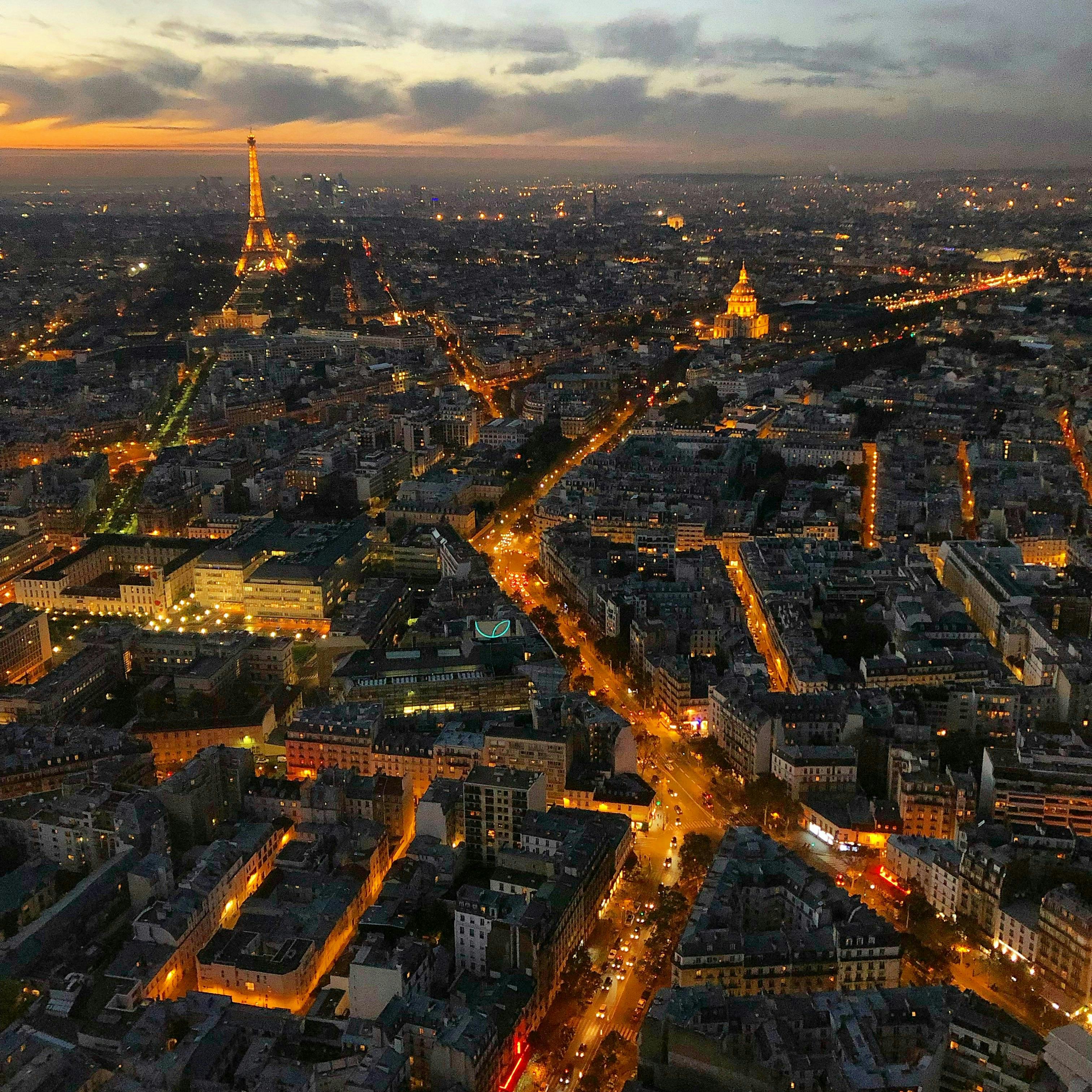 Aerial view of Paris at dusk with illuminated streets, the Eiffel Tower, and Les Invalides dome visible.