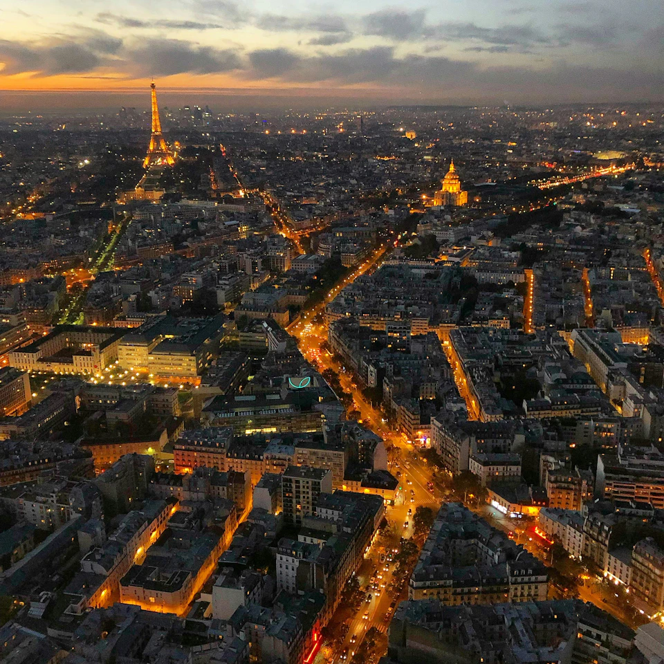 Aerial view of Paris at dusk with illuminated streets, the Eiffel Tower, and Les Invalides dome visible.
