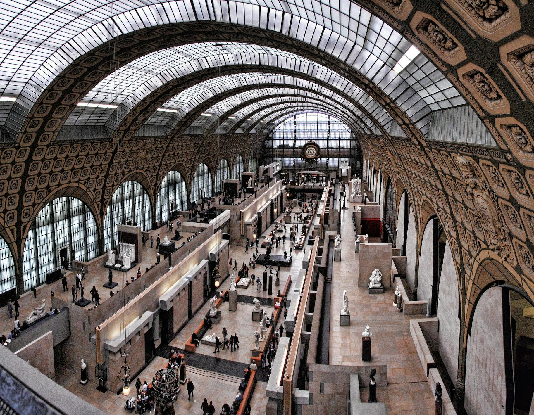 Interior of a large, arched museum hall with visitors walking among sculptures and exhibits. Large windows and ornate ceiling.