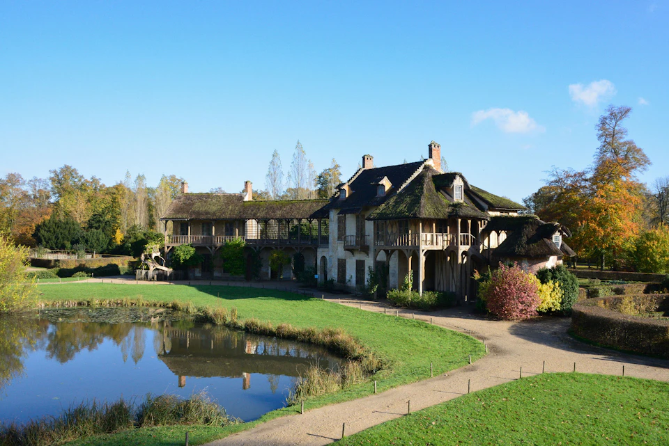 A quaint building with a thatched roof next to a pond, surrounded by greenery and trees under a clear blue sky.