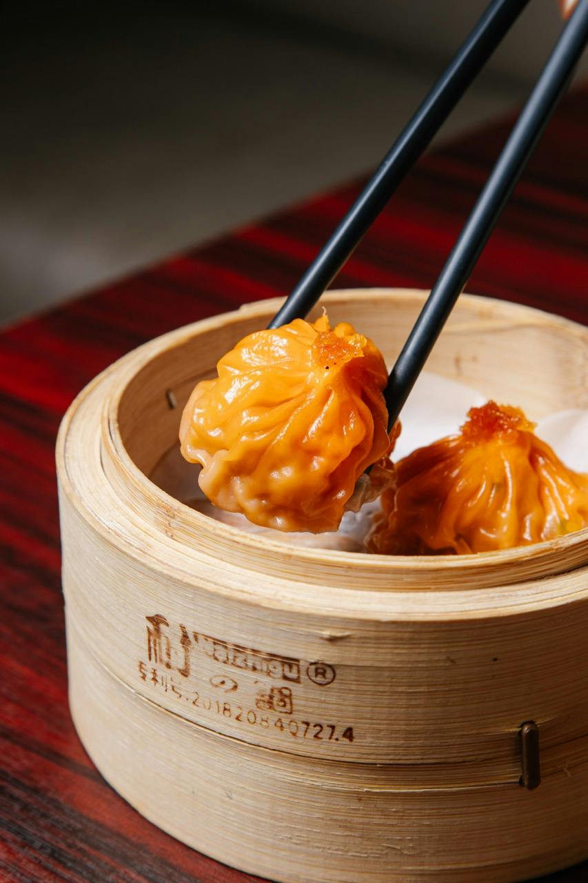 Chopsticks holding a steamed dumpling over a bamboo basket with another dumpling inside, set on a red wooden table.