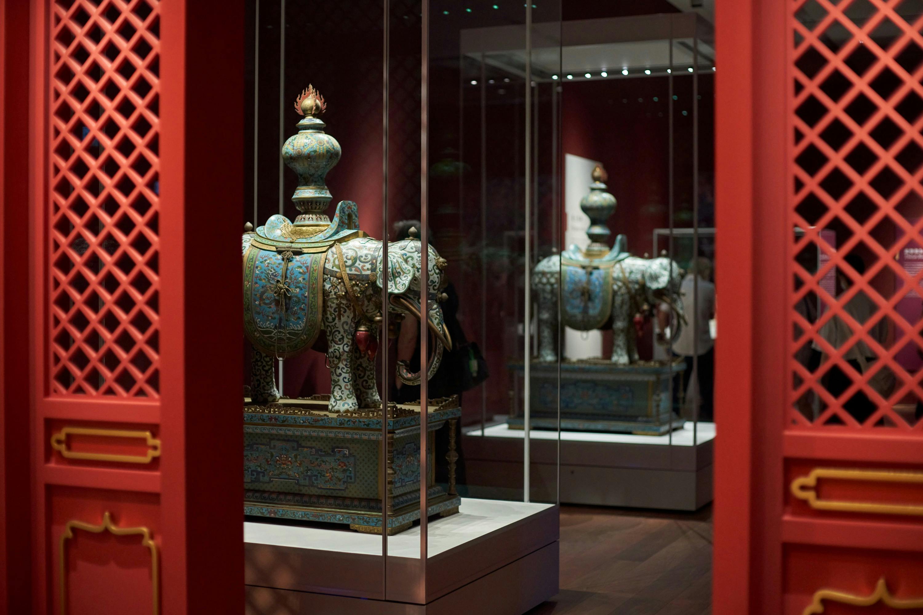 An ornate decorative elephant figurine displayed in a museum exhibit with mirrored panels and red walls.