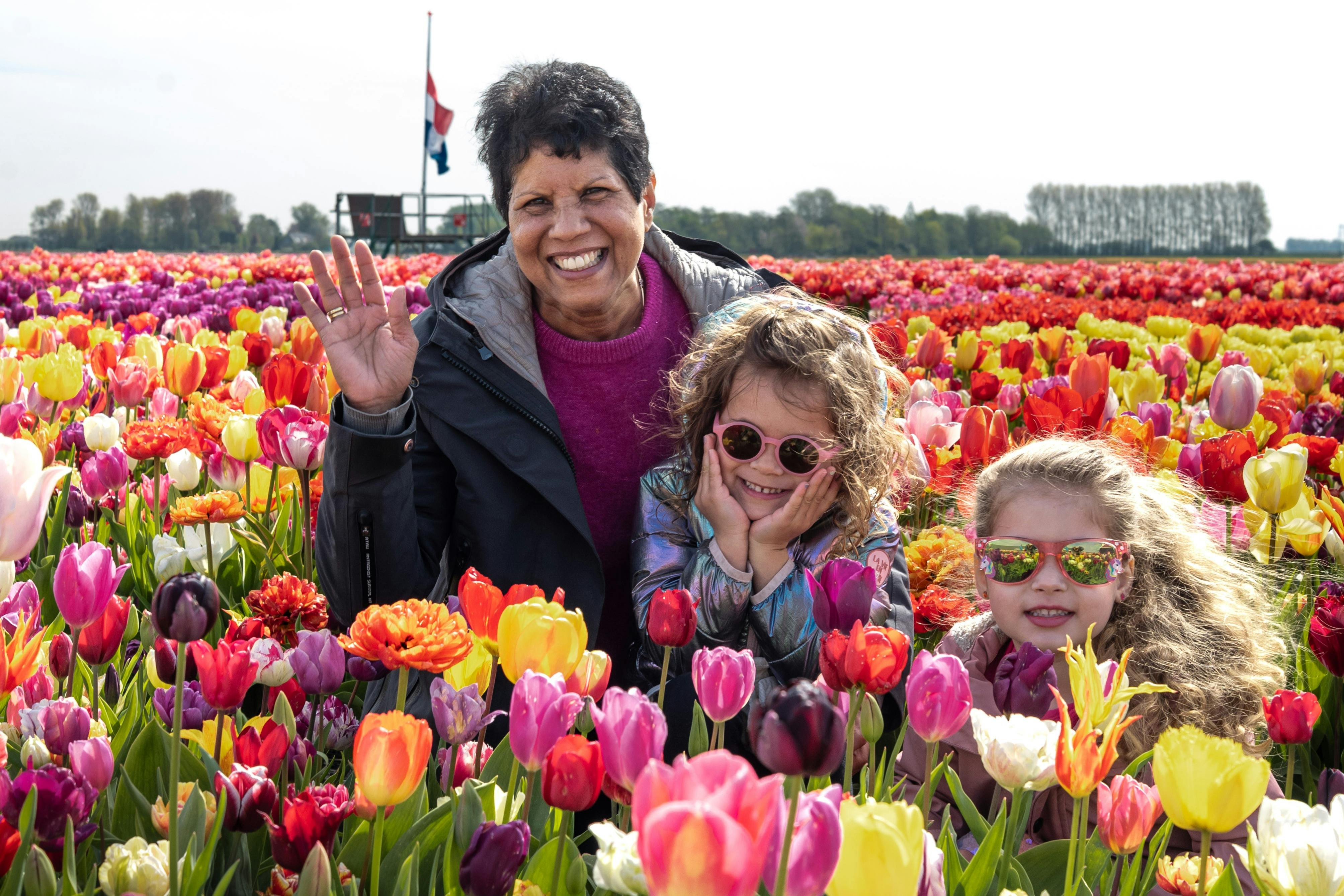 Three people smiling among colorful tulip fields under a bright sky, with trees and a flag in the background.