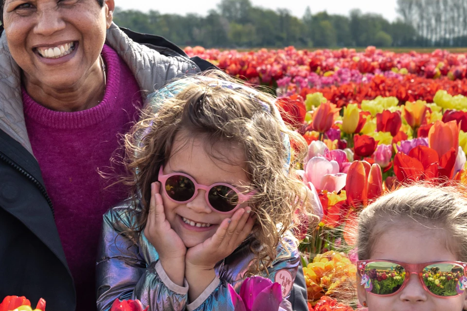 Three people smiling among colorful tulip fields under a bright sky, with trees and a flag in the background.