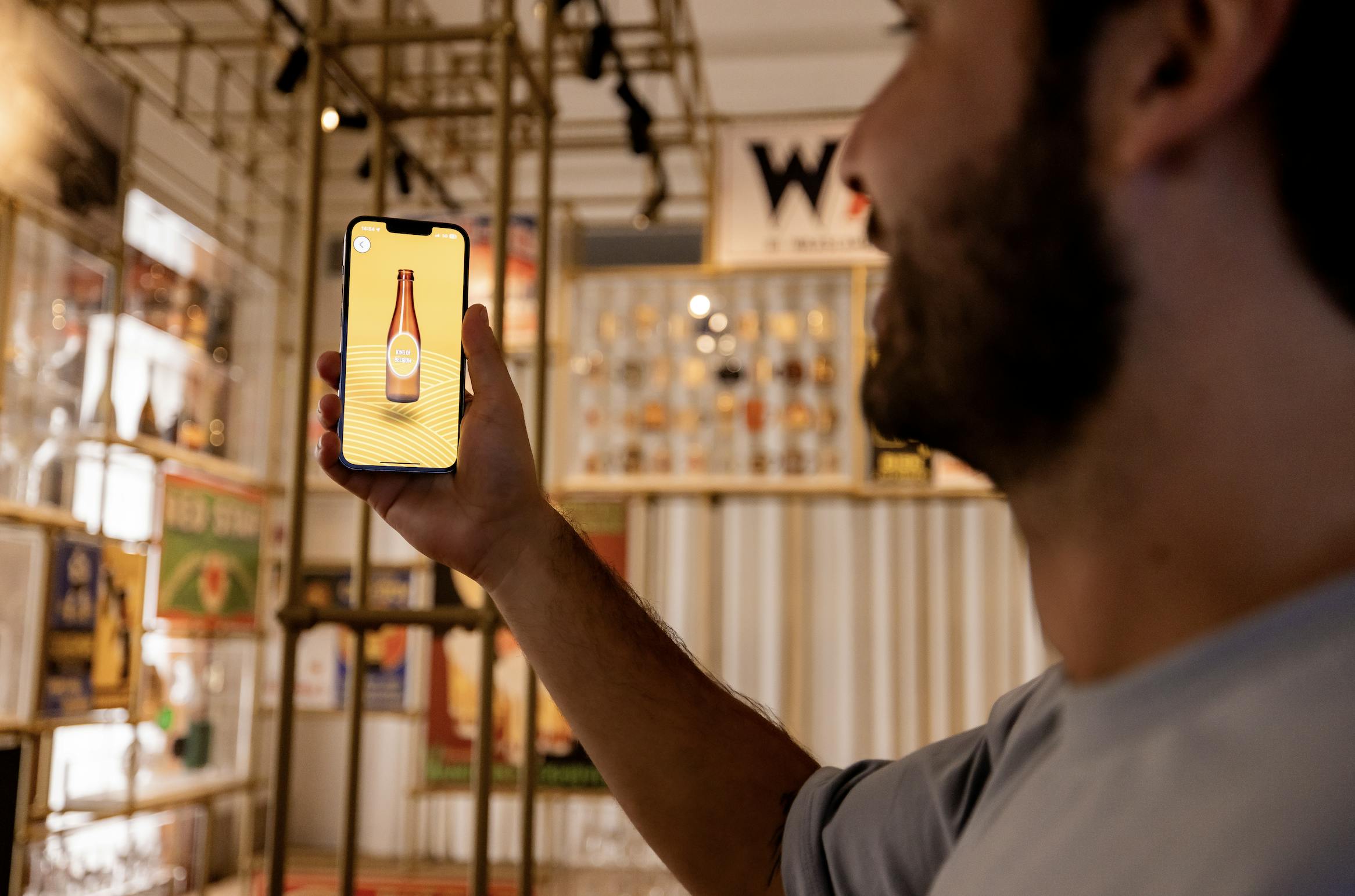 Un uomo tiene in mano uno smartphone che mostra l'immagine digitale di una bottiglia di birra, mentre si trova in uno spazio poco illuminato e pieno di esposizioni e cimeli vintage.