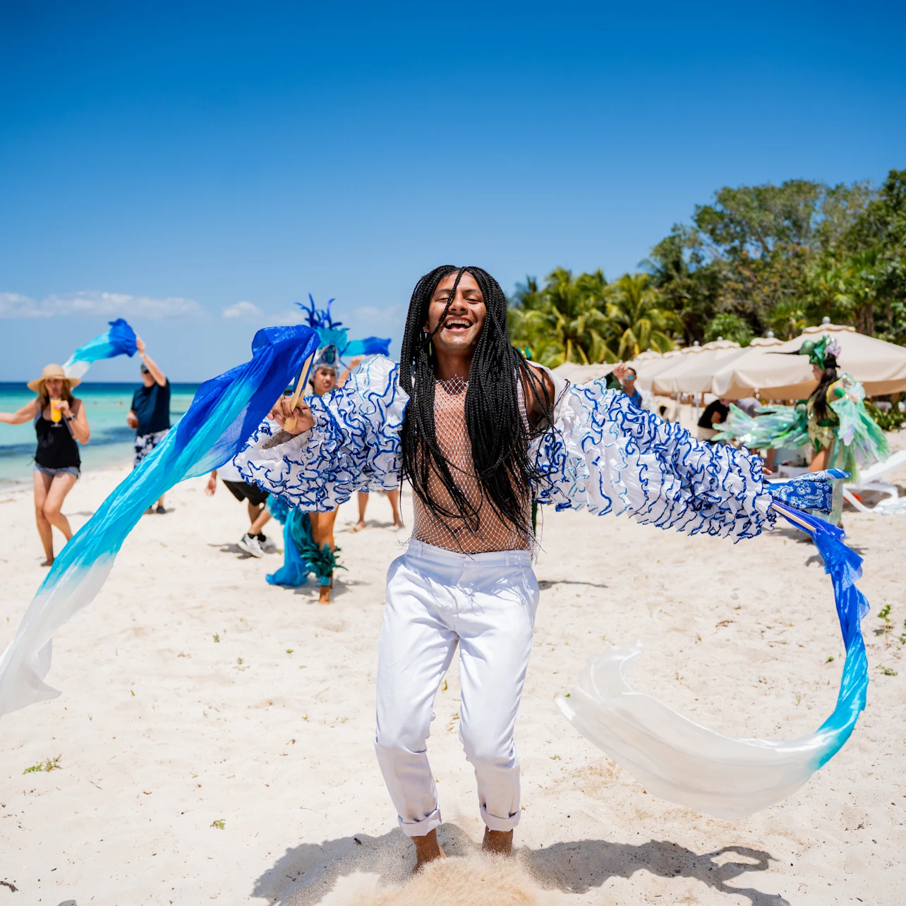 A person joyfully dances on a sandy beach under a clear blue sky, holding blue and white fabric strips, with others in the background.