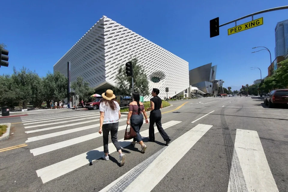 Three people crossing a street at a crosswalk, with a modern white building and traffic light visible in the background.