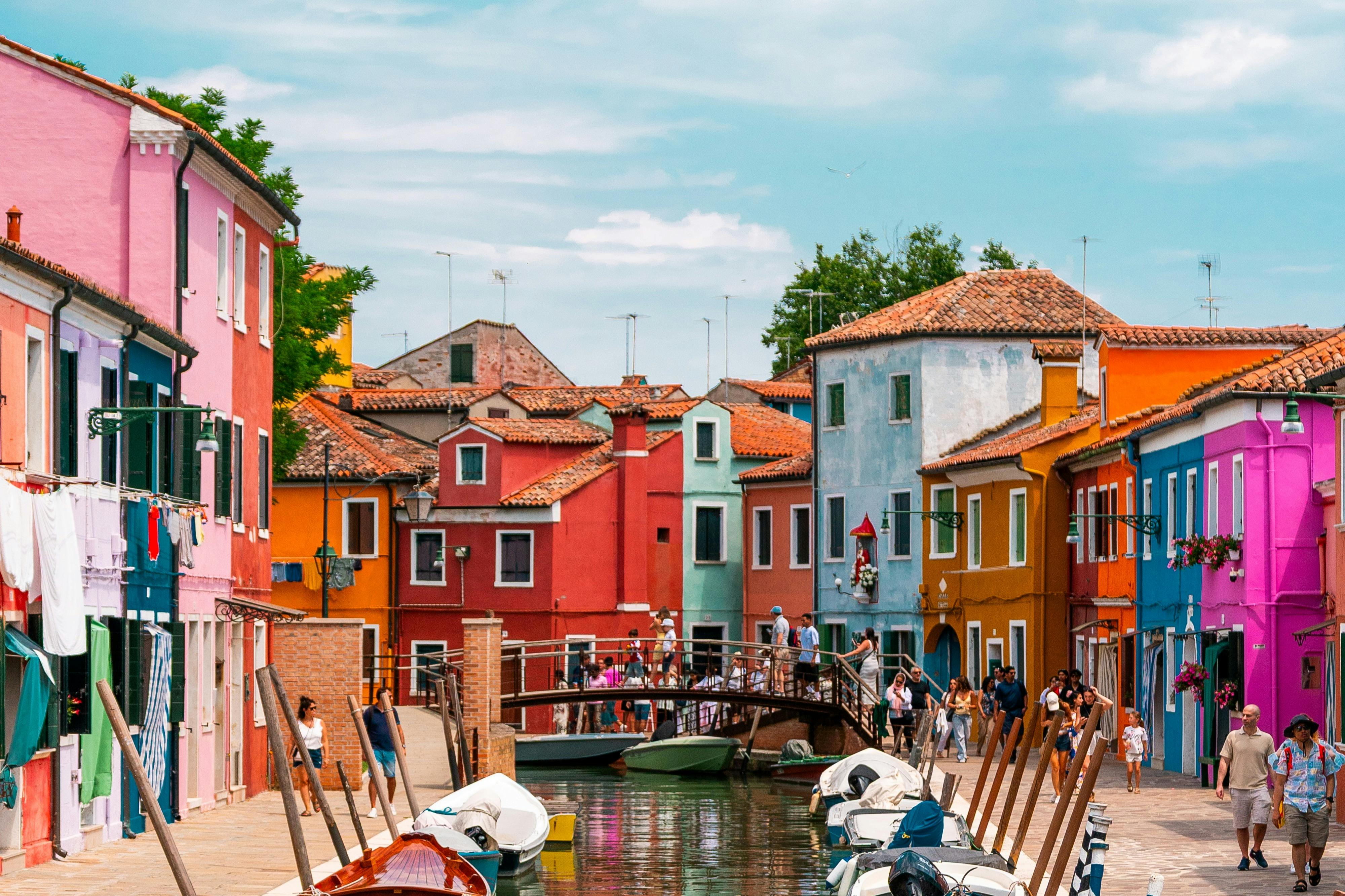 Um cenário de canal vibrante com casas coloridas, uma ponte pedonal, barcos atracados e pessoas a passear sob um céu azul.