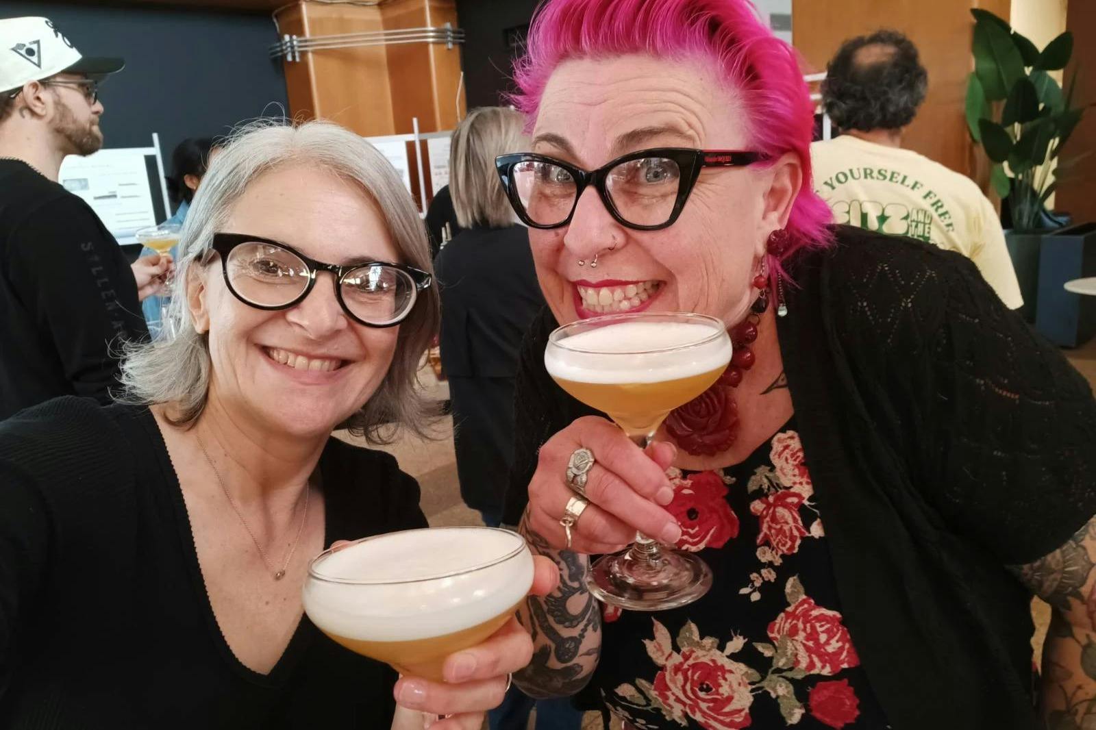 Two people smiling and holding cocktail glasses at a social event, with others and a display board visible in the background.