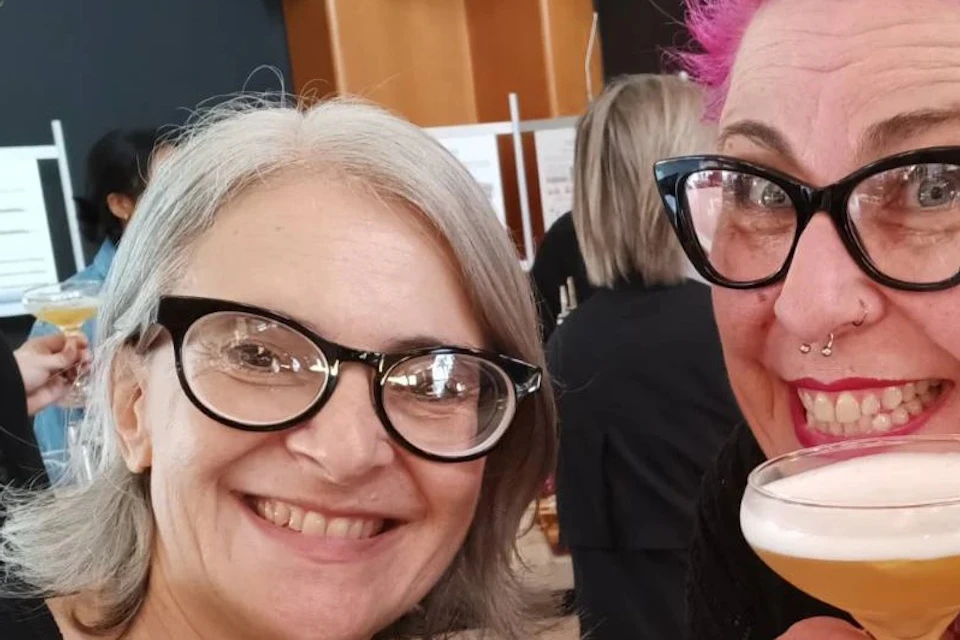 Two people smiling and holding cocktail glasses at a social event, with others and a display board visible in the background.