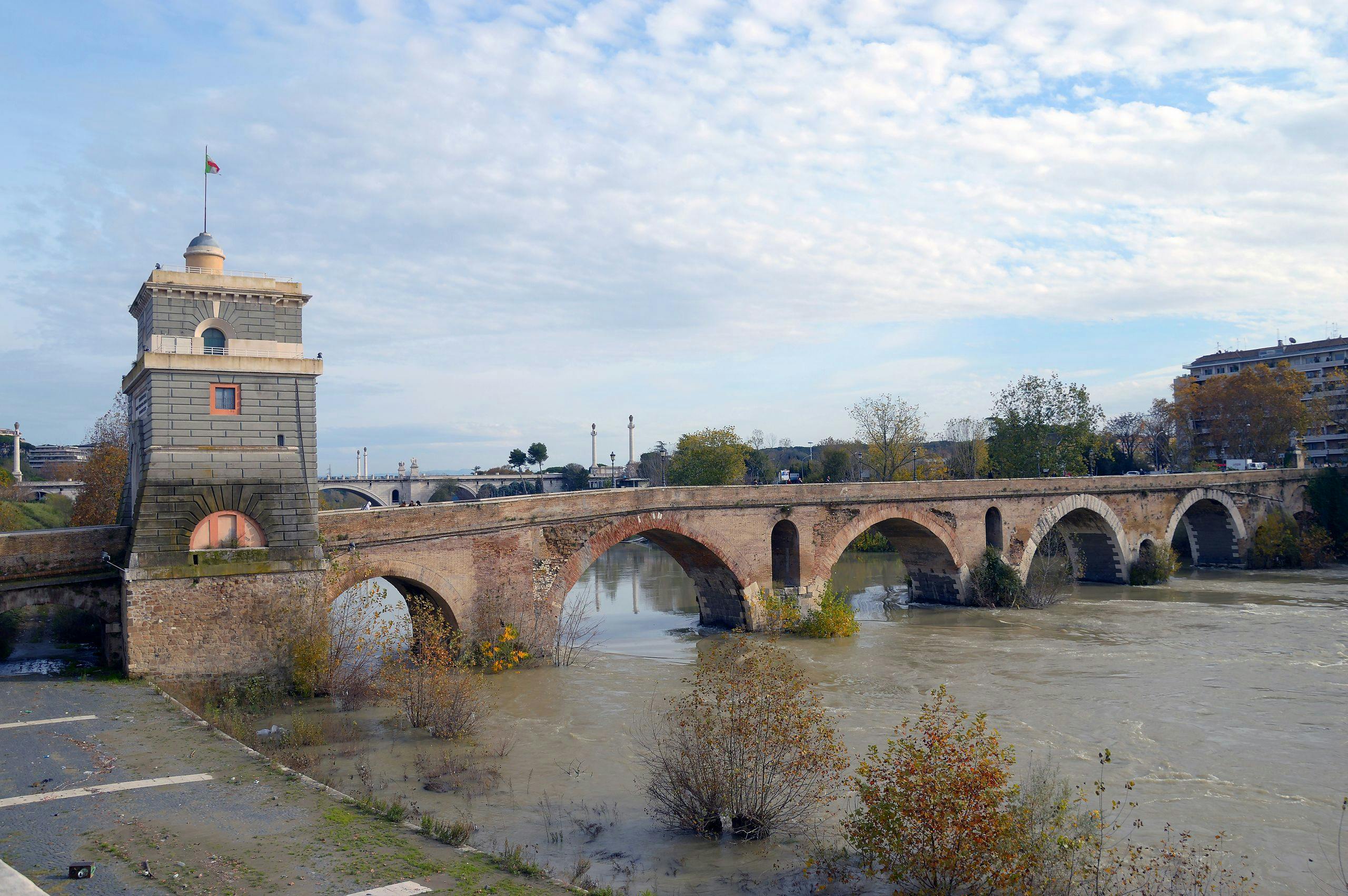 A stone bridge with multiple arches spans a river, with a tower on one end, under a partly cloudy sky. Trees and buildings in the background.