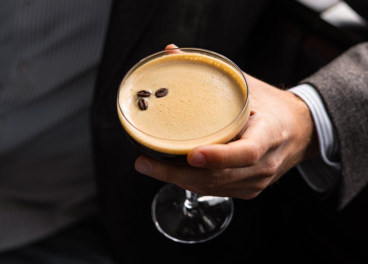 Ruka držící sklenici martini obsahující pěnivé espresso martini, ozdobené třemi kávovými zrny.
