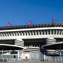 Modern stadions exteriör med röda strukturella accenter, cirkulärt överhäng, flera grindar och klarblå himmel ovanför.