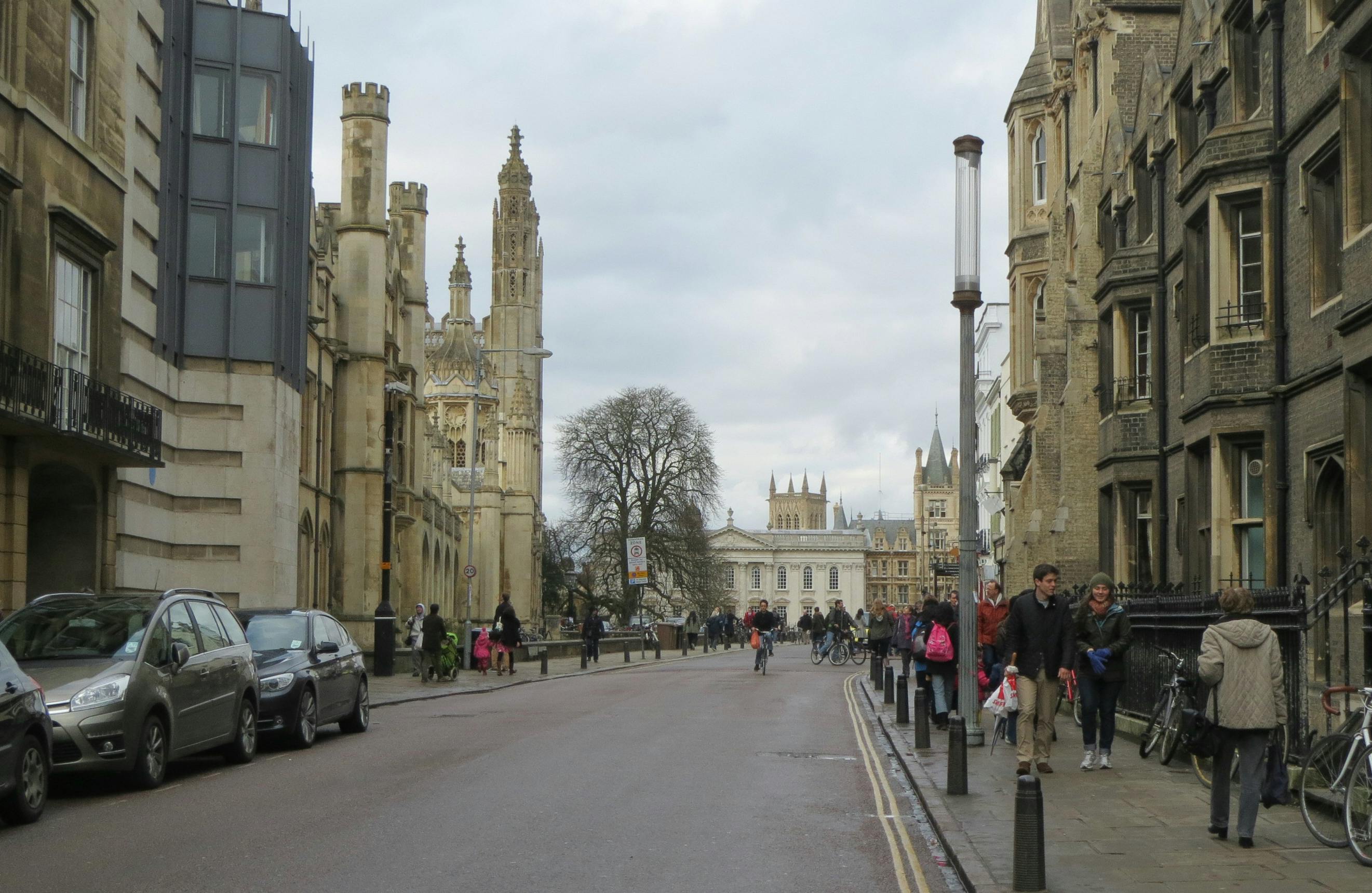 En gade med historiske stenbygninger, gående mennesker og cyklister. En stor kirke og et træ er synlige i baggrunden.