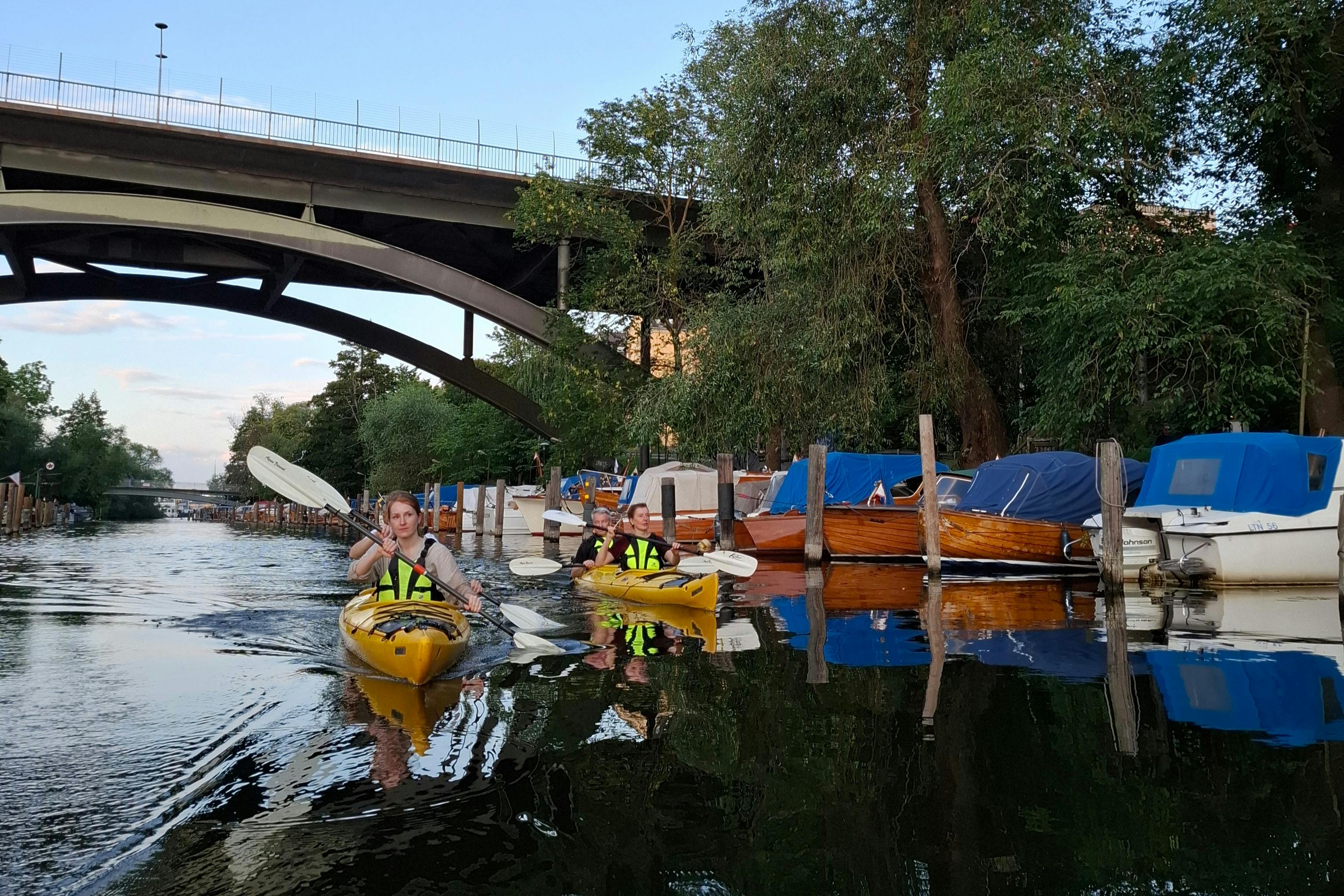 Deux personnes dans des kayaks jaunes descendent un canal sous un pont, avec des arbres et des bateaux le long de la voie d'eau.