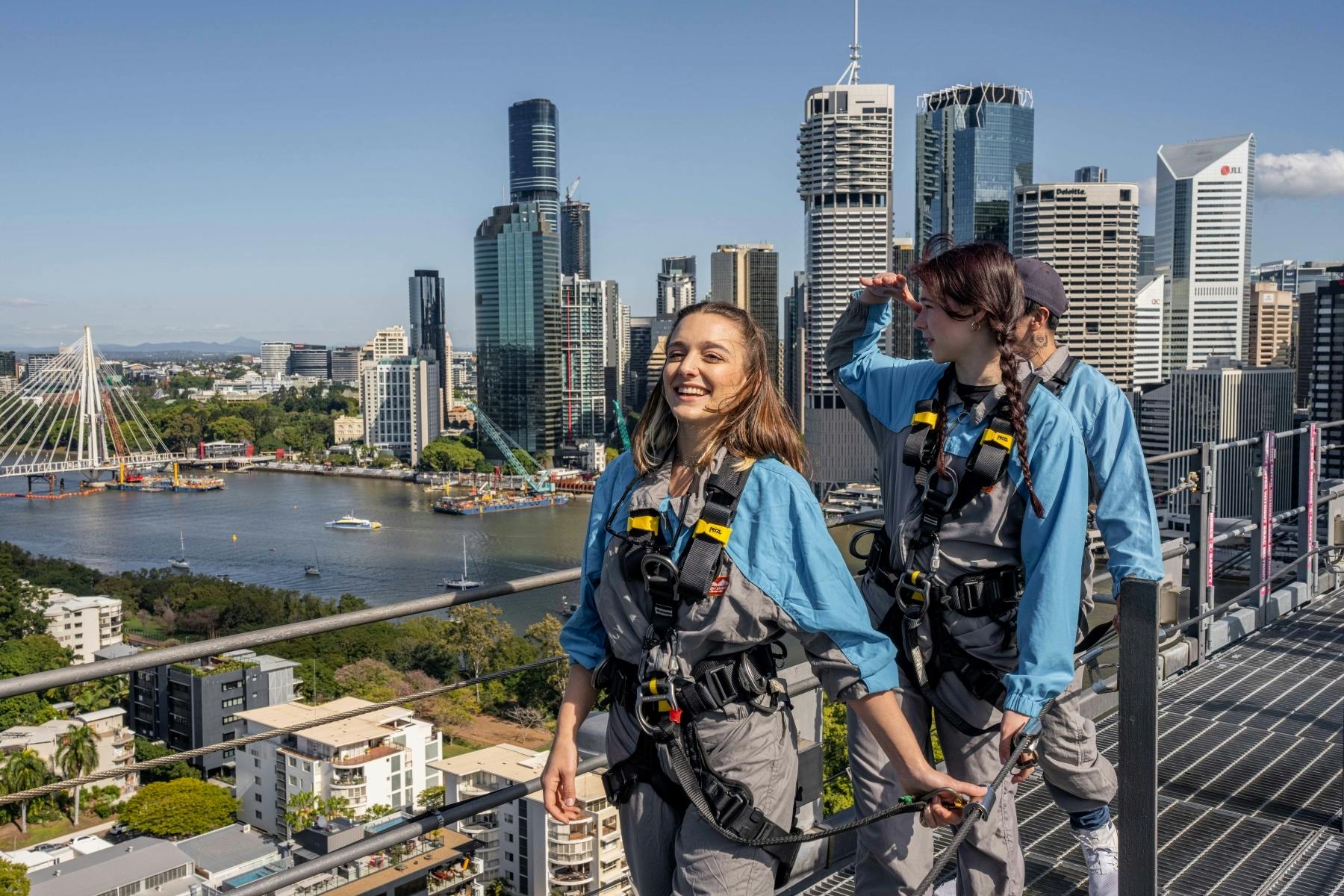 三个穿着安全装备的人在城市屋顶上，后面有摩天大楼和一条河流。 一个人微笑，另一个人遮住眼睛。