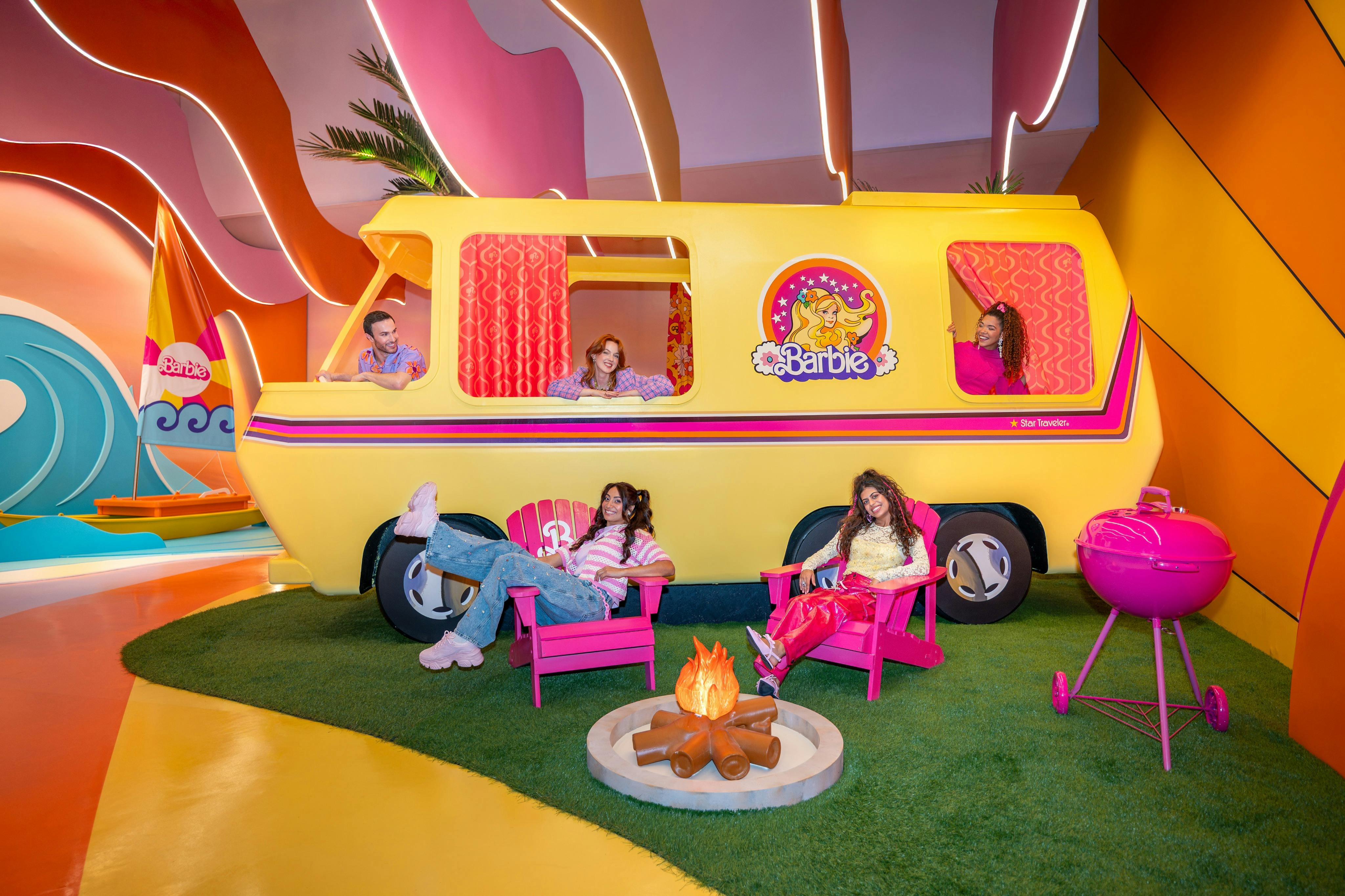 Čtyři lidé si užívají barbíny se žlutým karavanem, růžovými židlemi, táborovým ohněm a růžovým grilem na zelené umělé trávě.