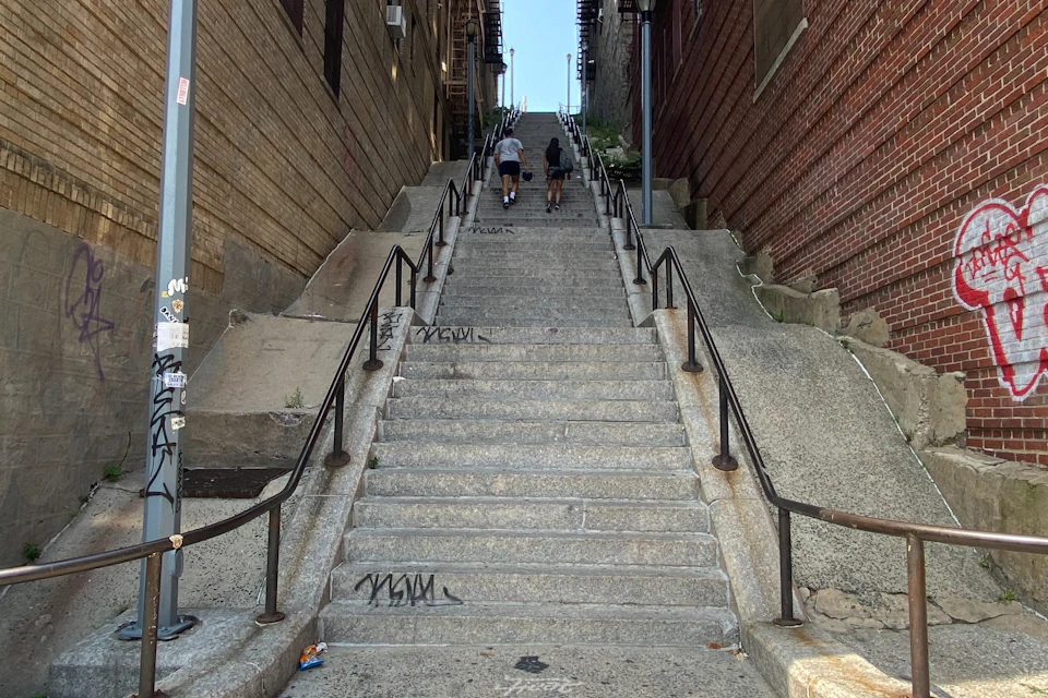 Two people climbing long concrete stairs with handrails on both sides, flanked by brick and wooden walls, under a clear sky.